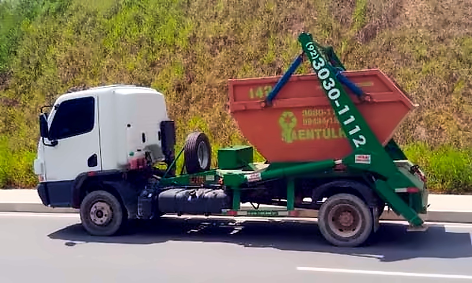 Caminhão poliguindaste verde carregando uma caçamba de entulho laranja da S.A. Entulho, parado no acostamento de uma via com encosta gramada ao fundo.